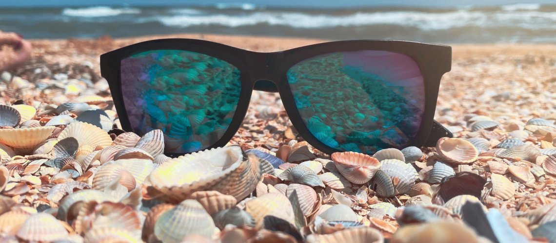 Sunglasses with blue lenses lie on a sandy shell beach on the sea front in sunny weather. Recreation and tourism concept.