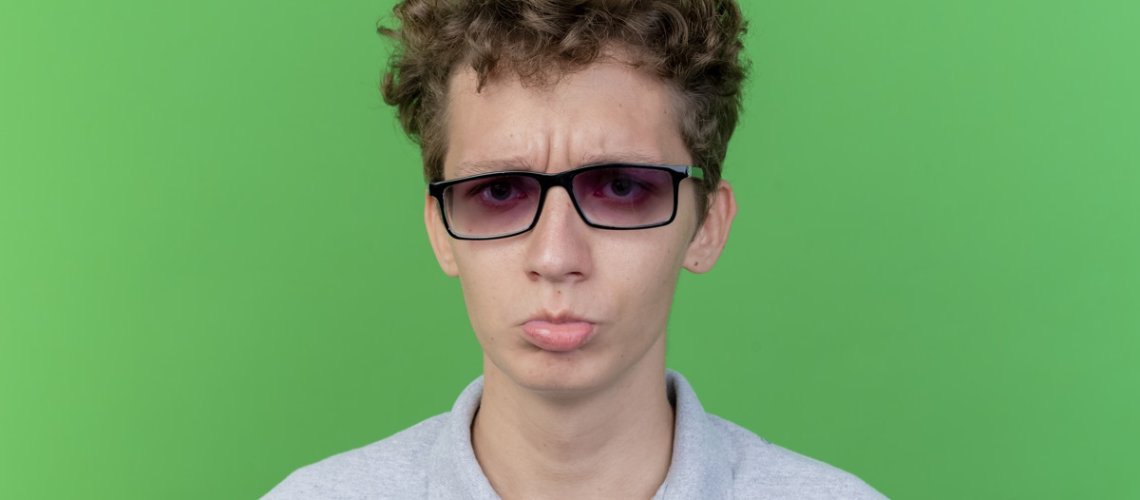 young man in black glasses wearing grey polo shirt looking at camera being displeased frowning standing over green background